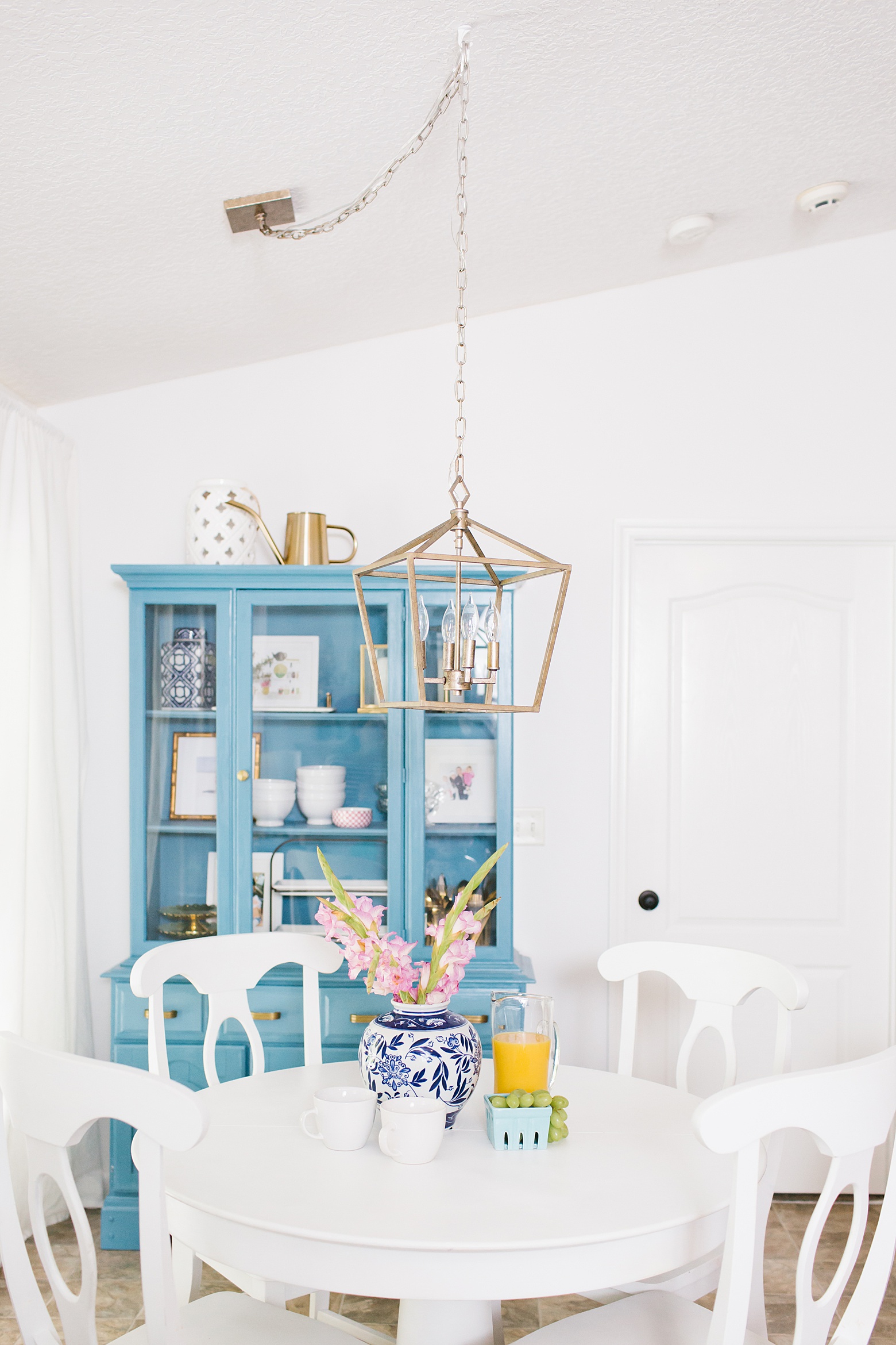 A bright white kitchen featuring a blue hutch, gold lantern pendant, white kitchen table, all white walls, white cabinets, and gold hardware on Megan Martin Creative